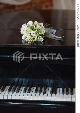 Wedding rings and white rose bouquet on black piano in rustic room with brick wall 127730774
