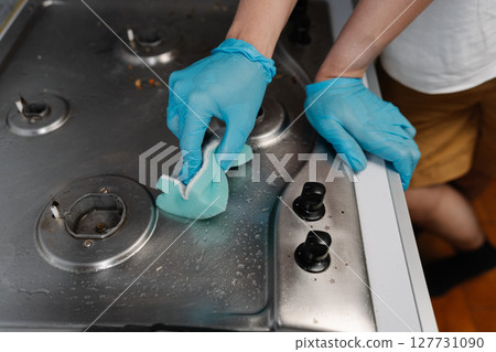 Cropped shot of housekeeper wearing blue gloves scrubbing dirty gas stove with sponge, applying cleaning solution for sparkling kitchen surfaces, close-up. Concept of cleanliness and housekeeping. 127731090