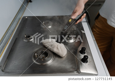 Top view of housewife cleaning dirty gas stove using steam cleaner and cloth, removing grease and burnt food residues, ensuring hygienic kitchen environment. Concept of modern housekeeping appliance. Top view of housewife cleaning dirty gas stove using steam cleaner and cloth, removing grease and burnt food residues, ensuring hygienic kitchen environment. Concept of modern housekeeping appliance. 127731145