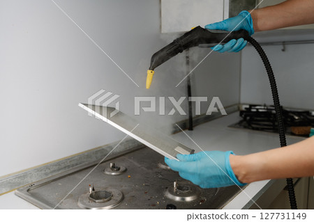 Cleaning company worker male wearing protective gloves sanitizing kitchen extractor filter using high-pressure steam cleaning equipment over gas stove. Concept of maintenance modern home appliance. 127731149