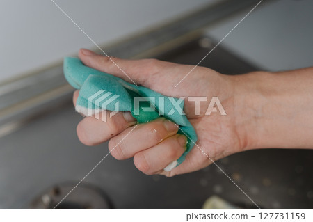 Housewife hand squeezing soapy water from sponge during cleaning dirty gas stove, to removing grime and grease, promoting hygiene and cleanliness in kitchen. Concept of domestic work and household. Housewife hand squeezing soapy water from sponge during cleaning dirty gas stove, to removing grime and grease, promoting hygiene and cleanliness in kitchen. Concept of domestic work and household. 127731159