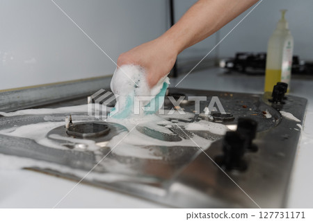Male hand scrubbing gas stove with sponge and detergent, promoting hygiene and achieving sparkling clean kitchen surface tackling dirt and grease. Concept of domestic work, housekeeping and household. Male hand scrubbing gas stove with sponge and detergent, promoting hygiene and achieving sparkling clean kitchen surface tackling dirt and grease. Concept of domestic work, housekeeping and household. 127731171