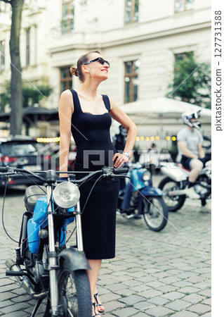 Portrait of pretty young adult stylish elegant woman wear black dress sit vintage retro blue motorcycle moped european city street Germany. Happy female person model enjoy having fun urban outside 127731388