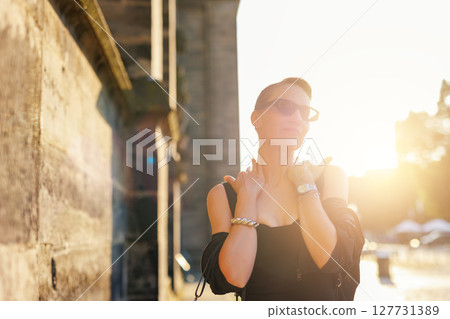 Portrait young adult elegant woman black dress and sunglasses smiles confidently in golden sunlight warm sunset backlit light against historic cathedral building. Female person walking city street Portrait young adult elegant woman black dress and sunglasses smiles confidently in golden sunlight warm sunset backlit light against historic cathedral building. Female person walking city street 127731389