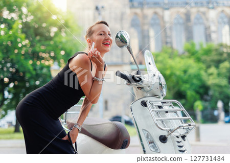 Portrait young adult beautiful woman looking in scooter bike mirror applying red lipstick against medieval cathedral Europe city street. Female person wear black dress heels makeup cosmetics outdoors 127731484