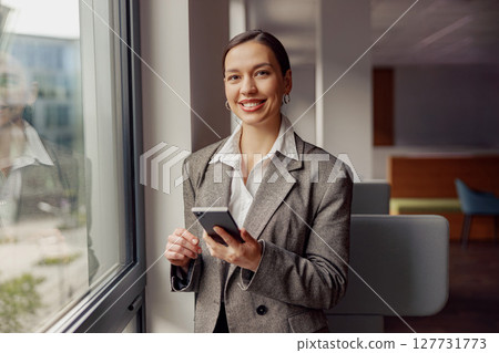 A Business Professional is Cheerfully Smiling by a Window while Holding a Smartphone in Their Hand A Business Professional is Cheerfully Smiling by a Window while Holding a Smartphone in Their Hand 127731773