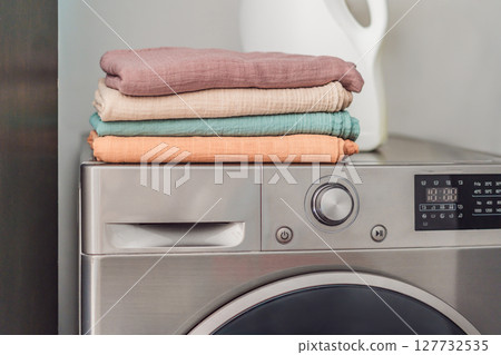 Stack of soft muslin baby swaddles neatly folded on a modern washing machine in a bright laundry room. Concept of baby care, hygiene, and gentle fabric for newborn essentials Stack of soft muslin baby swaddles neatly folded on a modern washing machine in a bright laundry room. Concept of baby care, hygiene, and gentle fabric for newborn essentials 127732535