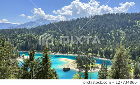 Mountain landscape with vibrant turquoise alpine lake surrounded by dense evergreen forest with distant mountain peaks in Sapiential region of Graubunden canton, Swiss 127734860
