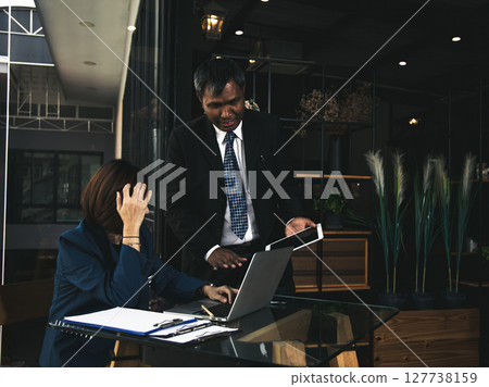 Businesswoman in navy suit sitting at glass desk with hand on forehead showing stress while businessman in black suit presenting data on tablet Businesswoman in navy suit sitting at glass desk with hand on forehead showing stress while businessman in black suit presenting data on tablet 127738159