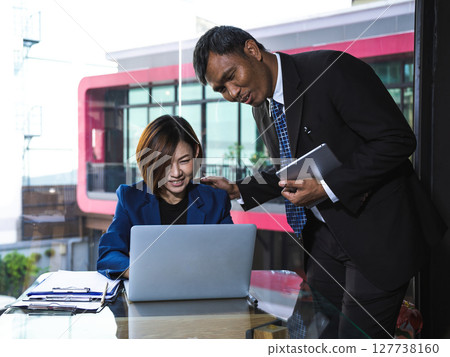 Businesswoman in navy suit smiling at laptop while businessman in black suit with tablet encourages and supports her by patting shoulder Businesswoman in navy suit smiling at laptop while businessman in black suit with tablet encourages and supports her by patting shoulder 127738160