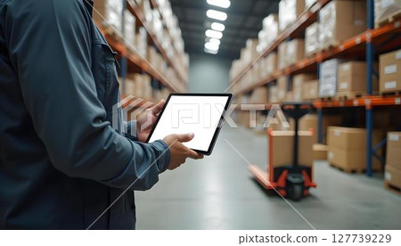 A man is focused on his work in a warehouse, holding a tablet amidst the rows of stored goods A man is focused on his work in a warehouse, holding a tablet amidst the rows of stored goods 127739229