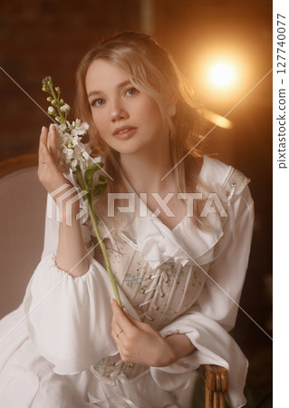 Young woman in white dress holding flower sitting on chair in dreamy warm-lit room 127740077