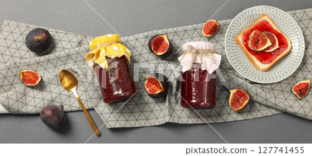 Figs, jars of jam, towel and bread with jam on plate on gray background, top view 127741455