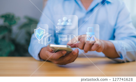 Person holding a smartphone interacts with a transparent overlay showing a login screen with security icons shield, padlock, user profiles. Online security, data protection, and user authentication. Person holding a smartphone interacts with a transparent overlay showing a login screen with security icons shield, padlock, user profiles. Online security, data protection, and user authentication. 127741628