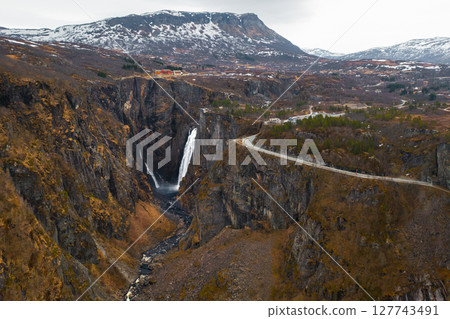 Magnificent Voringsfossen waterfall plunges into deep rugged canyon flanked by rocky cliffs. Winding road and sparse vegetation complete landscape 127743491