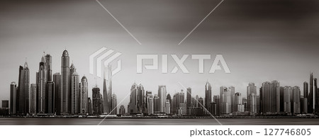 Monochrome city skyline with tall skyscrapers reflecting on calm water under a cloudy sky, Dubai Marina bay UAE 127746805