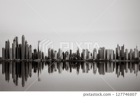 Monochrome city skyline with tall skyscrapers reflecting on calm water under a cloudy sky, Dubai Marina bay UAE 127746807