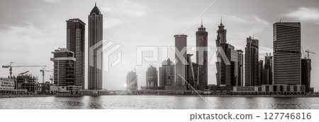 Monochrome city skyline with tall skyscrapers reflecting on calm water under a cloudy sky, Dubai Marina bay UAE 127746816