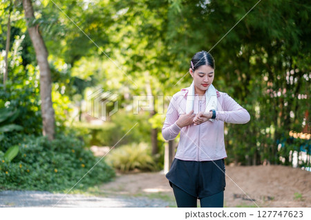 A woman in athletic wear checks her smartwatch while exercising in a vibrant, green park environment. 127747623