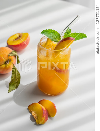 Peach drink or tea with ice, mint in a glass on a white table with fresh fruits and sunlight. 127751124