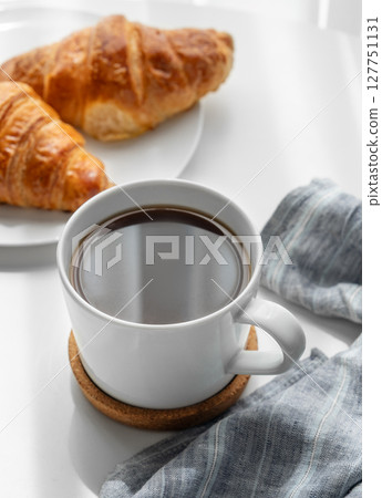 Coffee espresso cup and fresh croissants on a plate on a white table with morning shadows. Coffee espresso cup and fresh croissants on a plate on a white table with morning shadows. 127751131