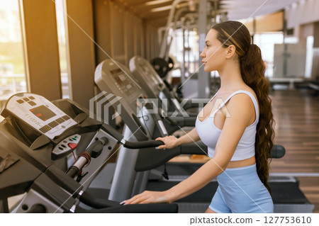 Side view of girl in sports bra doing heavy cardio workout on treadmill. Sexy muscular woman with slim body running in empty gym. Sport, bodybuilding concept. Side view of girl in sports bra doing heavy cardio workout on treadmill. Sexy muscular woman with slim body running in empty gym. Sport, bodybuilding concept. 127753610
