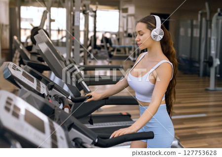 Young sporty woman running on machine in the gym and listening music in headphones 127753628