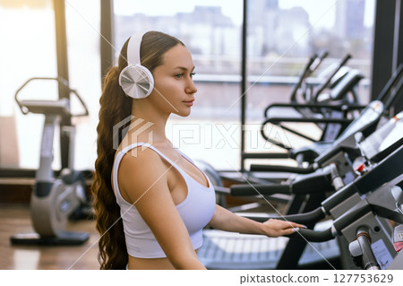 Young sporty woman running on machine in the gym and listening music in headphones Young sporty woman running on machine in the gym and listening music in headphones 127753629