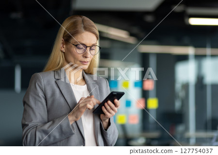 Mature business woman in office environment using smartphone for communication and management tasks. She appears confident and focused, showcasing leadership and professionalism. Mature business woman in office environment using smartphone for communication and management tasks. She appears confident and focused, showcasing leadership and professionalism. 127754037