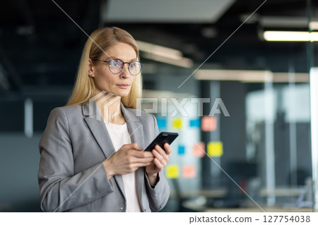 Mature businesswoman in office holding smartphone, embodying leadership and professionalism. Gray suit and glasses highlight focus and confidence in digital communication and business management. Mature businesswoman in office holding smartphone, embodying leadership and professionalism. Gray suit and glasses highlight focus and confidence in digital communication and business management. 127754038