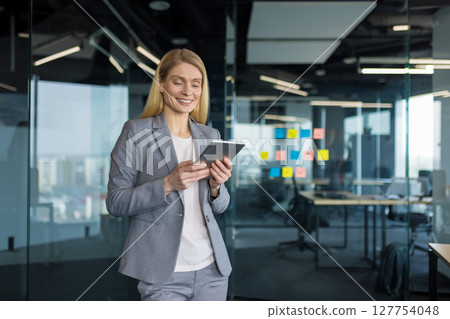 Successful woman in business suit standing inside office with tablet computer in hands smiling happily. Happy businesswoman reading electronic report and viewing investment data online. 127754048