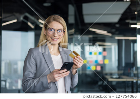 Successful joyful woman standing inside the office with a phone in her hands and a bank credit card. Businesswoman looking at the camera with satisfaction, having made a successful money transfer. 127754066
