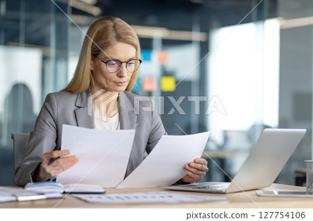 A professional woman reviews documents at her desk, using a laptop in a modern office setting. Focus on the business and work. 127754106