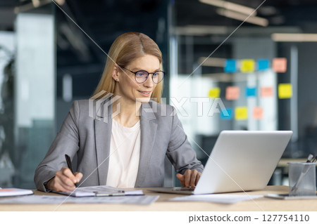 Attractive blonde businesswoman is working with laptop, taking notes. She is in a modern office with a glass wall, indoor. Attractive blonde businesswoman is working with laptop, taking notes. She is in a modern office with a glass wall, indoor. 127754110