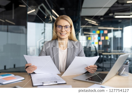 A businesswoman is analyzing documents at her desk with a laptop and smiling in a modern office setting, showcasing her focus and expertise. 127754127