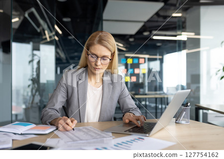 A businesswoman in a modern office setting is focused while working on a laptop and reviewing documents at her desk, analyzing data. 127754162