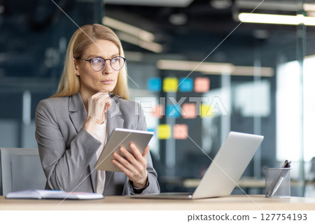 Thoughtful businesswoman in glasses reviews information on a tablet, seated at a desk with a laptop. She is in an office. 127754193