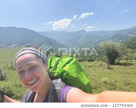 Hiking tourism adventure. Backpacker hiker woman taking selfie picture. Hiker girl lady tourist with backpack walking near mountain. Young happy woman enjoy hike trekking tourism active vacation 127754364