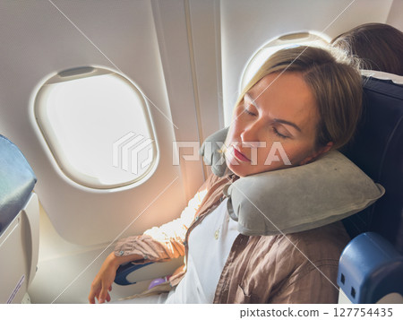 Rest on plane. Young woman napping sitting by porthole in business jet using neck pillow for travel. Passenger girl flying in plane. Girl sleeping in airplane going on trip vacation. Traveling girl Rest on plane. Young woman napping sitting by porthole in business jet using neck pillow for travel. Passenger girl flying in plane. Girl sleeping in airplane going on trip vacation. Traveling girl 127754435