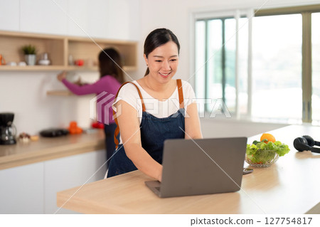 Remote Cooking and Wellness. Woman working on a laptop while another prepares fresh ingredients in the kitchen. Remote Cooking and Wellness. Woman working on a laptop while another prepares fresh ingredients in the kitchen. 127754817