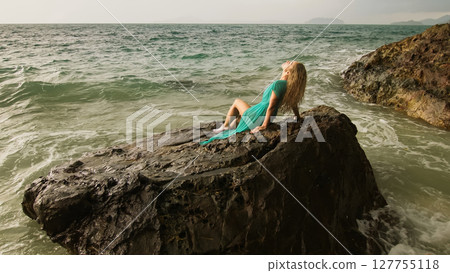 Woman sitting on rock of sea reef stone, stormy cloudy ocean. Blue swimsuit dress tunic. Concept resort coastline tourism summer holidays 127755118
