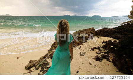 Woman walks to the stormy cloudy ocean on sand beach. Girl in blue swimsuit dress tunic. Concept rest in sea, tropical resort coastline tourism Woman walks to the stormy cloudy ocean on sand beach. Girl in blue swimsuit dress tunic. Concept rest in sea, tropical resort coastline tourism 127755194