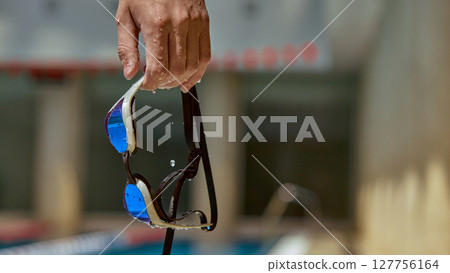 Wet swimsuit goggles held by hand near pool edge with blurred background 127756164