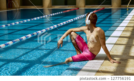 Man relaxing poolside in indoor swimming facility wearing headphones and vibrant pink swim shorts 127756191