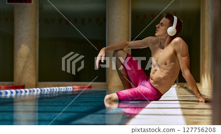 Young man relaxing poolside in swim gear with headphones 127756208