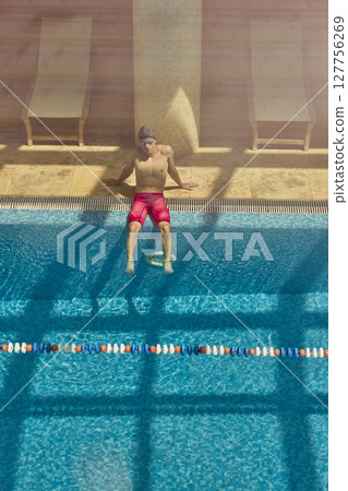 Man relaxing at edge of indoor swimming pool in red shorts with sunlit reflections on water Man relaxing at edge of indoor swimming pool in red shorts with sunlit reflections on water 127756269