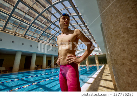 Swimmer standing confidently by indoor pool under arched glass roof with hands on hips Swimmer standing confidently by indoor pool under arched glass roof with hands on hips 127756285