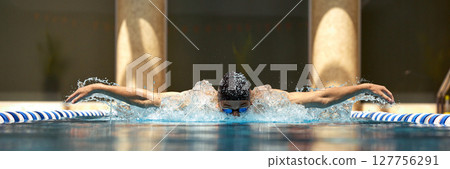 Swimmer performing butterfly stroke in indoor pool with determination and focus Swimmer performing butterfly stroke in indoor pool with determination and focus 127756291