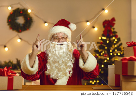 Santa sitting at desk in workshop, looking at camera during video call and sharing interesting ideas Santa sitting at desk in workshop, looking at camera during video call and sharing interesting ideas 127756548