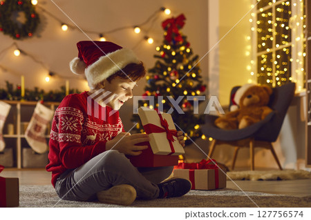 Happy boy in Santa hat opening his Christmas present sitting in the living-room at night Happy boy in Santa hat opening his Christmas present sitting in the living-room at night 127756574